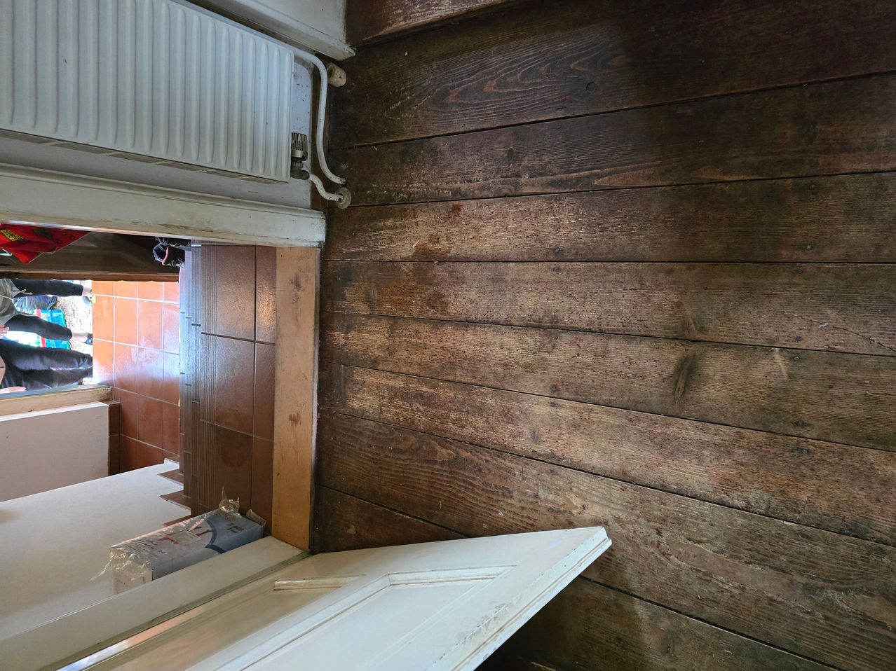 Eine Holzwand, wahrscheinlich Teil eines Hauses, weist sichtbare Abnutzungserscheinungen auf. Die Wand besteht aus Holzbrettern, von denen einige Flecken und Markierungen aufweisen. Unten im Bild befindet sich eine weiße Tür mit weißem Rahmen.
