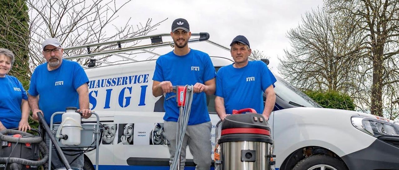 Zwei Männer in blauen Shirts und Mützen stehen neben einem Van mit der Aufschrift 'Service GI'. Einer hält einen roten Staubsauger, der andere einen schwarzen. Dahinter steht ein Baum.