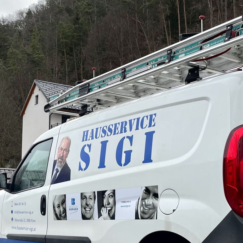 Ein weißer Van mit Dachgepäckträger steht vor einem Haus. Seitlich sind blaue Buchstaben zu lesen, die Hausservice Sigi bedeuten.