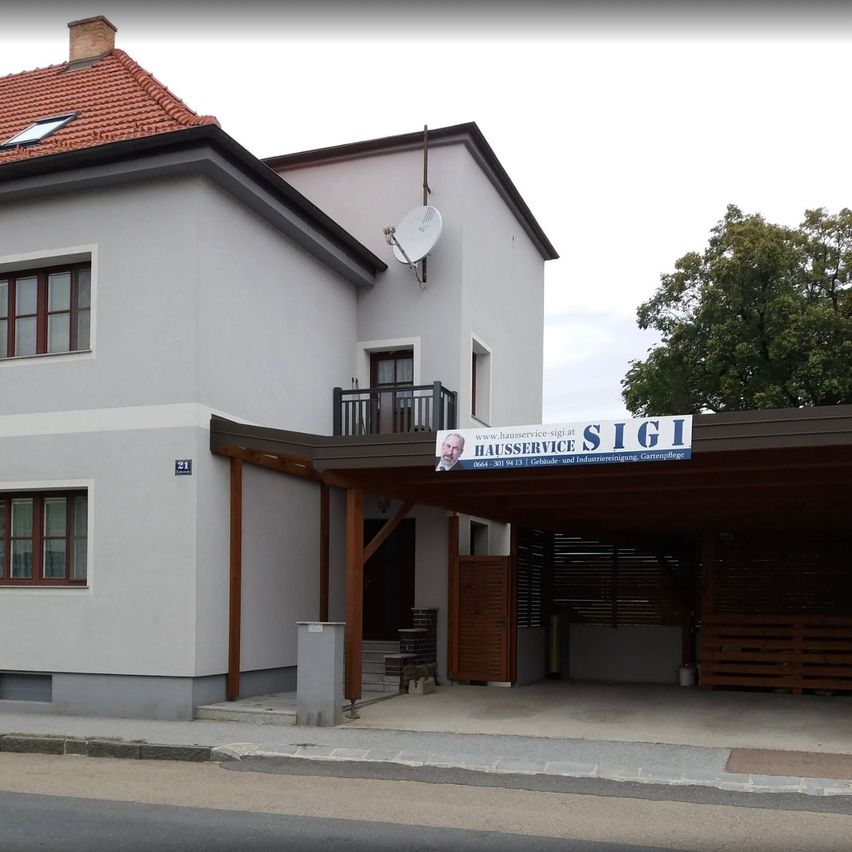 Ein weißes Gebäude mit rotem Dach, Satellitenschüssel und einem Schild mit der Aufschrift 'Hausservice SIGI'. Es hat einen Balkon, mehrere Fenster und eine Einfahrt mit Holzläden.