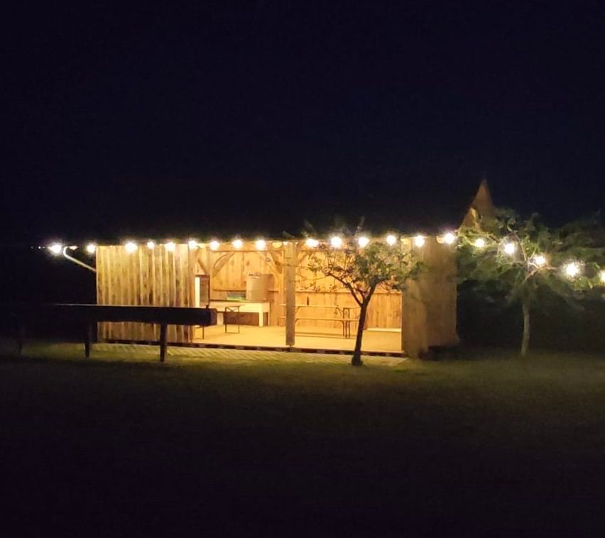 Ein von Lichterketten beleuchteter Holzschuppen bei Nacht, mit einer kleinen Bühne und Bänken davor.