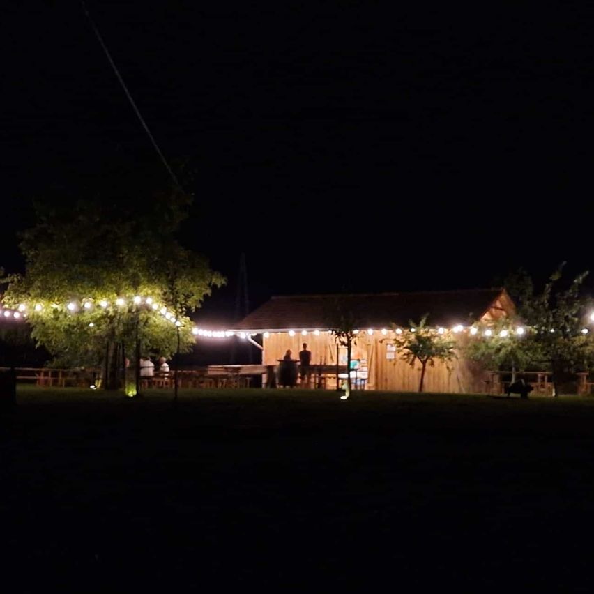 A rustic wooden building is illuminated by string lights at night. People are gathered around tables outside, enjoying the warm glow. Trees and a dark sky complete the scene.