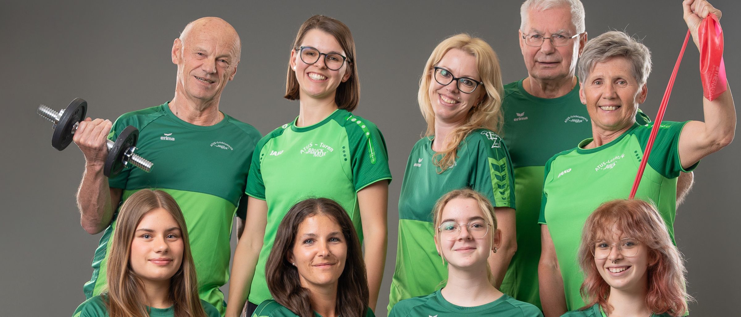 Eine Gruppe von Menschen in grünen T-Shirts und schwarzen Leggings posiert für ein Foto. Einige von ihnen halten Hanteln und ein rotes Band. Der Mann mit dem grauen Bart hält eine Hantel. Eine Frau mit Brille hält ein rotes Band.