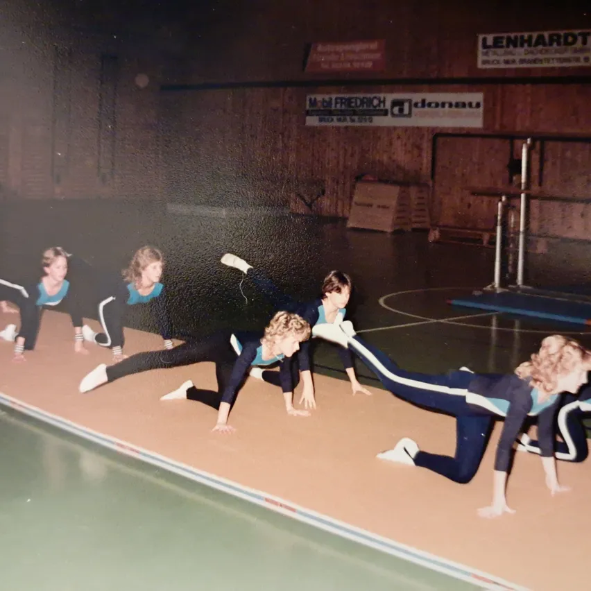 Eine Gruppe junger Frauen macht Gymnastik auf einer Matte in einer Turnhalle.