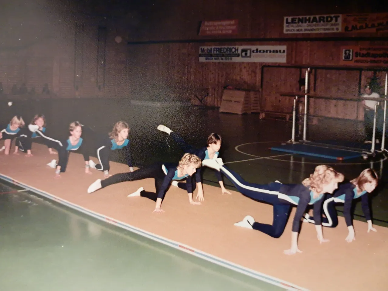 Eine Gruppe junger Frauen macht Gymnastik auf einer Matte in einer Turnhalle.