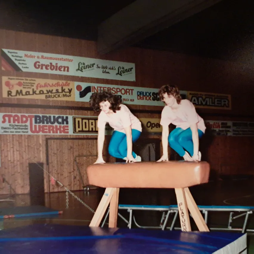 Zwei Frauen führen eine Balanceübung auf einer Holzplattform in einer Turnhalle durch. Sie tragen passende blaue Outfits.