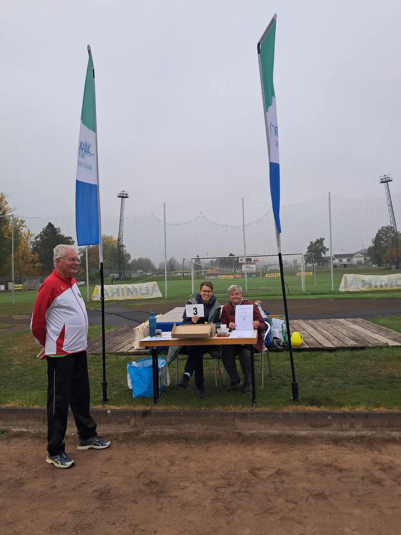 Drei Personen stehen auf einem Sportplatz, eine Person steht vor einem Tisch mit der Nummer 3.