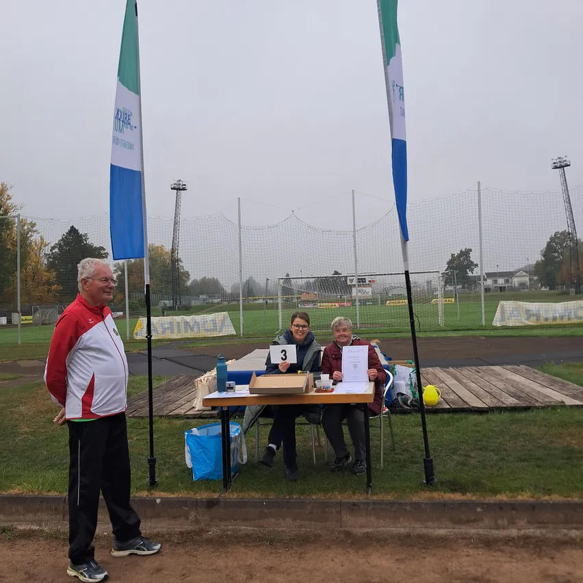 Drei Personen stehen auf einem Sportplatz, eine Person steht vor einem Tisch mit der Nummer 3.