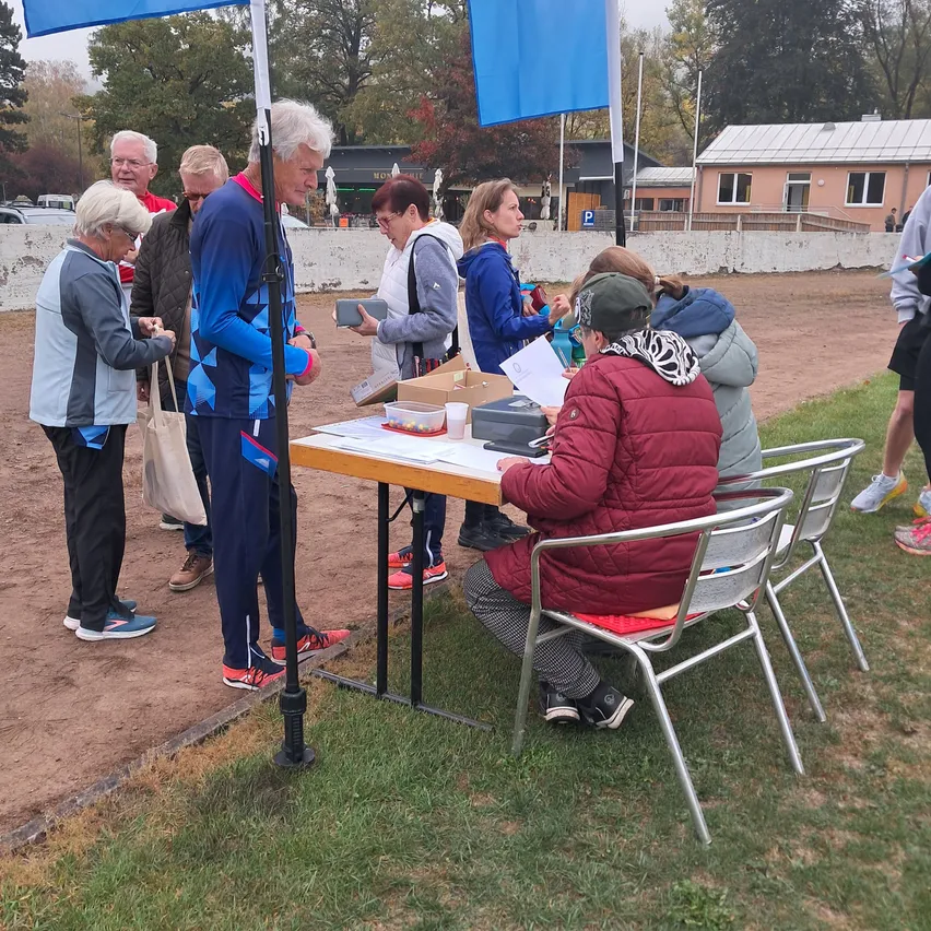Eine Gruppe von Menschen versammelt sich um einen Tisch im Freien. Eine Frau sitzt und schreibt auf Papier, während andere um sie herum stehen. Im Hintergrund befinden sich Fahnen und ein Gebäude.