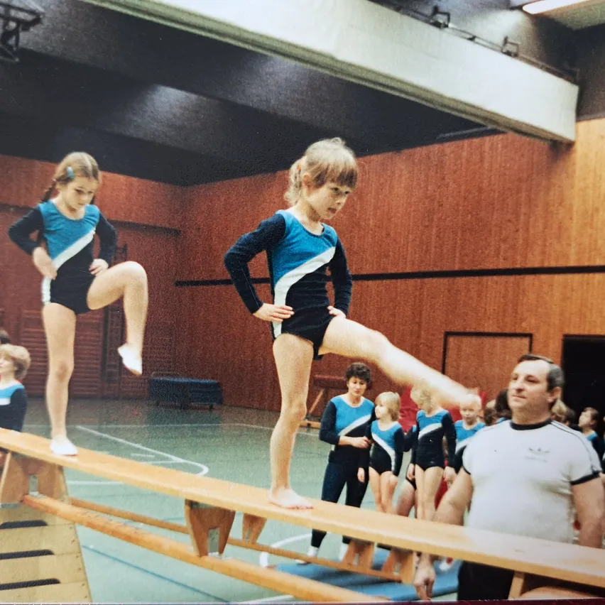 Zwei junge Mädchen führen Gymnastik auf einer Balancestange in einer Turnhalle aus. Ein Mädchen steht auf der Stange, während das andere springt. Ein Mann beobachtet sie von der Seite.