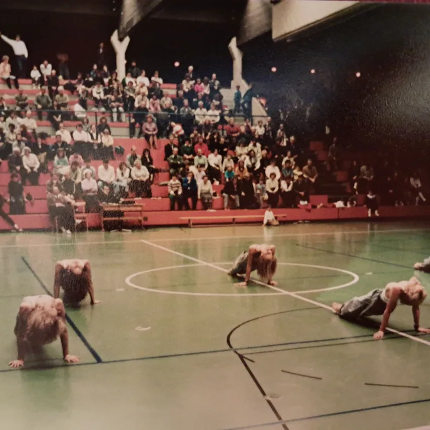 Eine Gruppe von Menschen führt eine synchronisierte Übungsroutine auf einem grünen Hallensportplatz durch. Das Publikum sitzt auf roten Tribünen im Hintergrund.