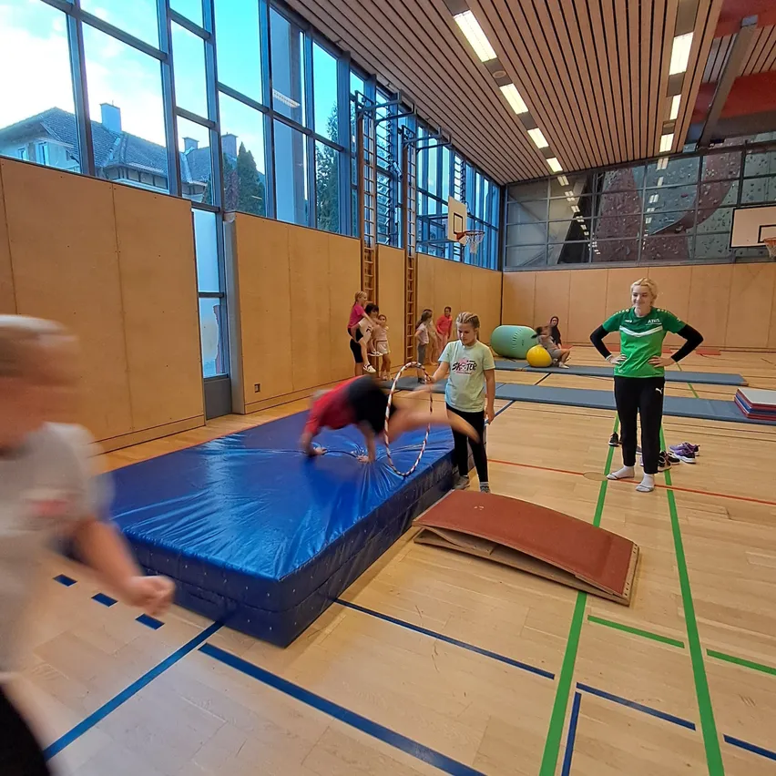 In einem Fitnessstudio sind eine Gruppe von Menschen mit Gymnastikaktivitäten beschäftigt. Eine Person macht einen Handstand auf einer blauen Matte, während eine andere Person einen Hula-Hoop hält.