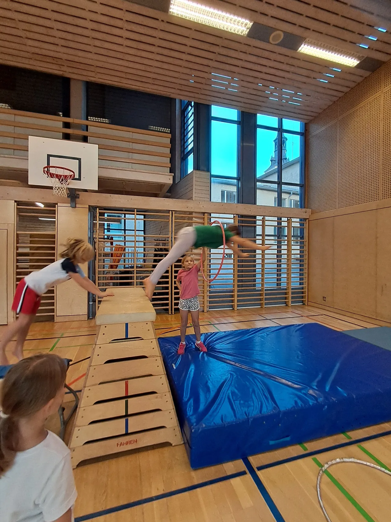 Eine Gruppe junger Mädchen spielt in einer Turnhalle. Ein Mädchen macht einen Handstand auf einer blauen Matte, während ein anderes Mädchen ihr hilft.