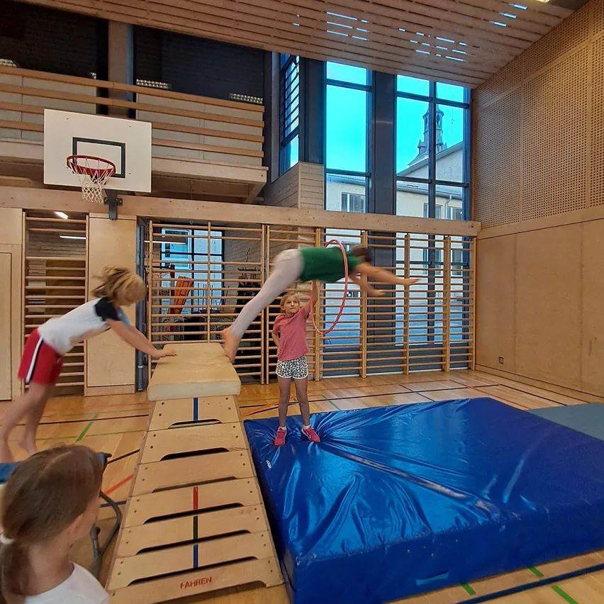 Eine Gruppe junger Mädchen spielt in einer Turnhalle. Ein Mädchen macht einen Handstand auf einer blauen Matte, während ein anderes Mädchen ihr hilft.