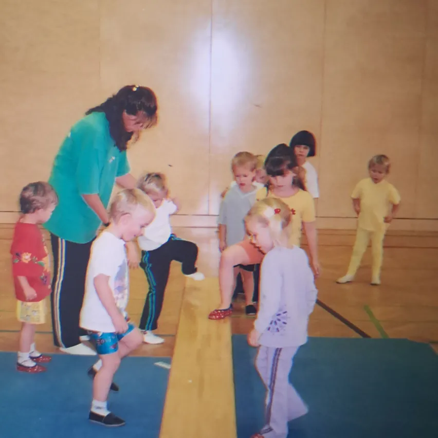 Eine Gruppe von Kindern und einer erwachsenen Frau sind in einem Fitnessstudio. Die Kinder stehen auf einem blauen Teppich und einem hölzernen Balancebalken.