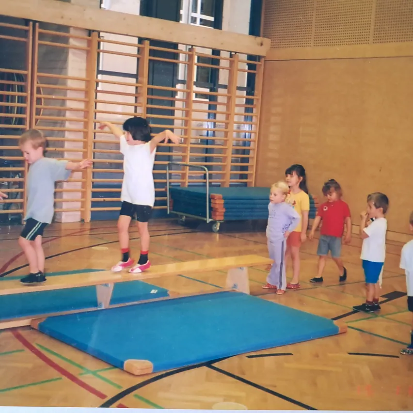 Eine Gruppe junger Kinder übt das Gleichgewicht auf einem Holzbalken in einer Turnhalle.