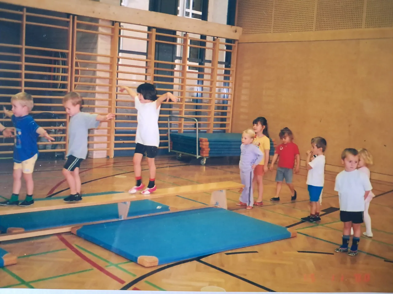 Eine Gruppe junger Kinder übt das Gleichgewicht auf einem Holzbalken in einer Turnhalle.