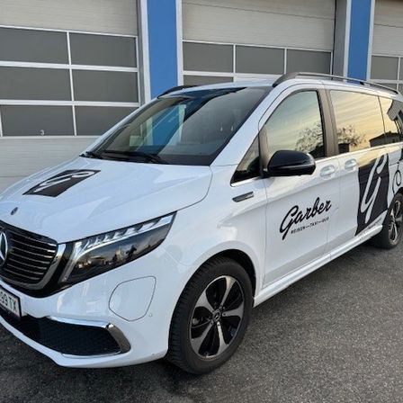 Ein weißer Mercedes-Benz V-Klasse Van mit schwarzen Akzenten und dem Wort Garber an der Seite ist vor einer Garage geparkt.