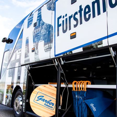 Die Seite eines Fürstenhof-Busses mit blau-weißer Lackierung, die das Wort 'Fürstenhof' deutlich zeigt. Im Inneren des Busses befinden sich blaue Regenschirme und ein Holzschild mit der Aufschrift 'Garber Exklusiv'.