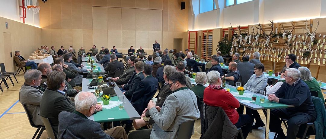Eine Konferenz findet in einem Raum mit Holzböden und großen Fenstern statt. Ein Mann steht an einem Podium und spricht zu einem sitzenden Publikum. Die Tische sind mit grünen Tüchern, Blumen und Gläsern mit Getränken gedeckt.