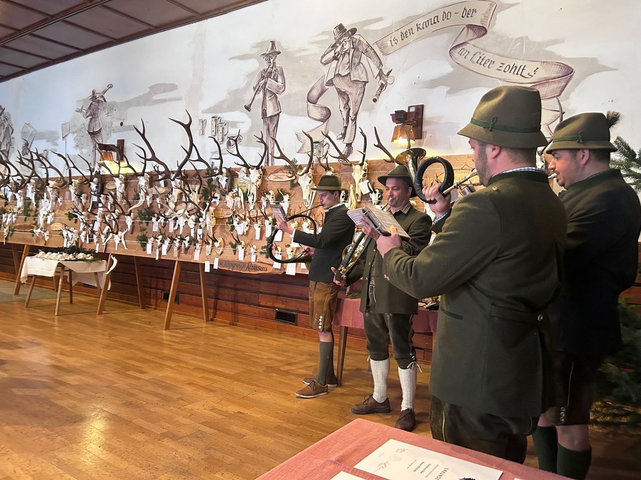 Drei Männer in traditioneller Kleidung spielen Instrumente in einem Raum mit Wandgemälde, Geweihen und Holzboden.