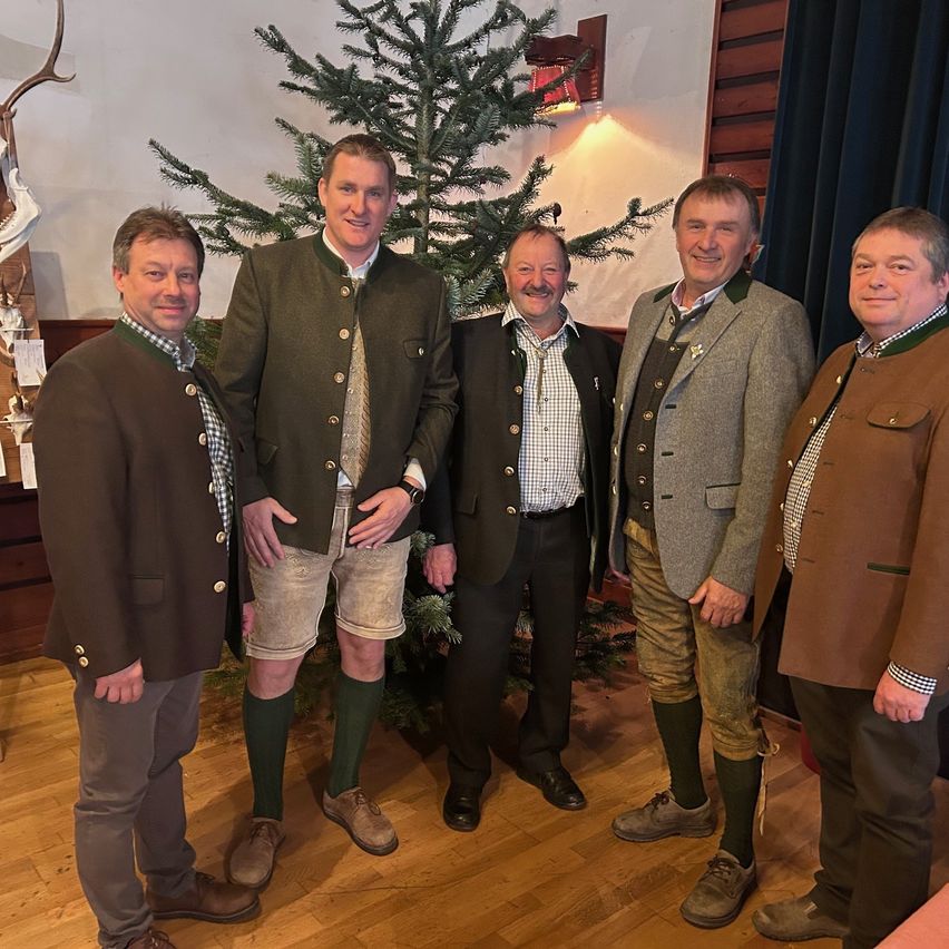 Fünf Männer in traditioneller deutscher Kleidung posieren vor einem geschmückten Weihnachtsbaum. Sie lächeln und scheinen sich bei einer festlichen Veranstaltung zu befinden.