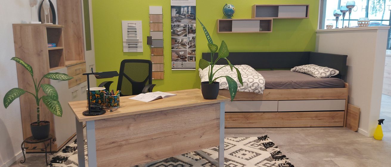 Ein Heimbüro mit Schreibtisch, Stuhl, Topfpflanze und Bett an einer grünen Wand.