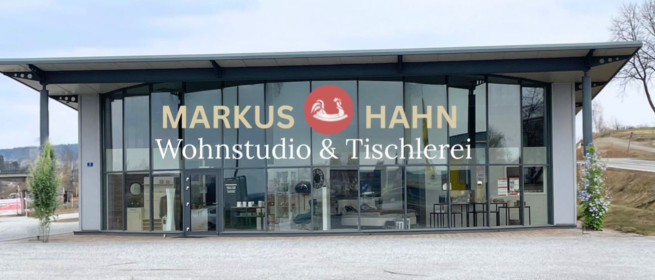 Außenansicht eines modernen Gebäudes mit Glaswänden, auf denen der Name 'Markus Hahn Wohnstudio & Tischlerei' steht. Im Inneren sind Möbel und Dekoration sichtbar.