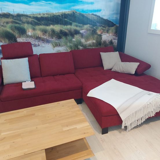 Ein modernes rotes Ecksofa mit Kissen, einer weißen Decke und einem hölzernen Couchtisch befindet sich in einem Raum mit einer Strandlandschaftstapete.