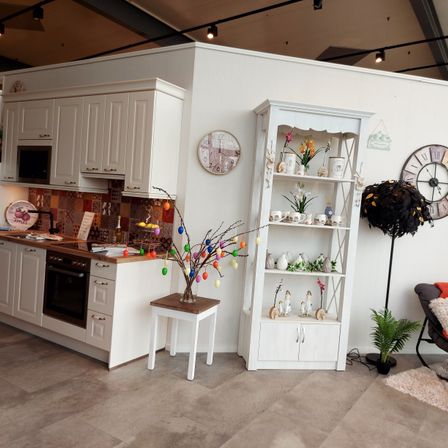 Eine weiße Küche mit Mikrowelle, Backofen und verschiedenen Regalen, geschmückt mit Blumen, Vasen und Dekorationsartikeln, vor einer weißen Wand mit Uhren.