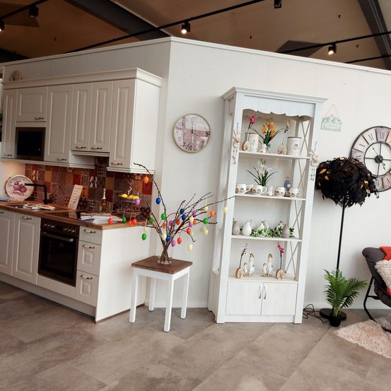 Eine weiße Küche mit Mikrowelle, Backofen und verschiedenen Regalen, geschmückt mit Blumen, Vasen und Dekorationsartikeln, vor einer weißen Wand mit Uhren.