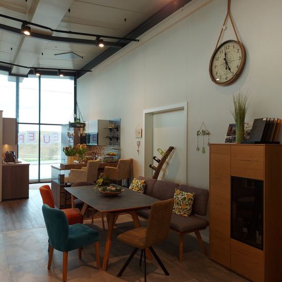 Ein Essbereich mit einem Holztisch und gepolsterten Stühlern, einer Küche im Hintergrund und einer Wanduhr.