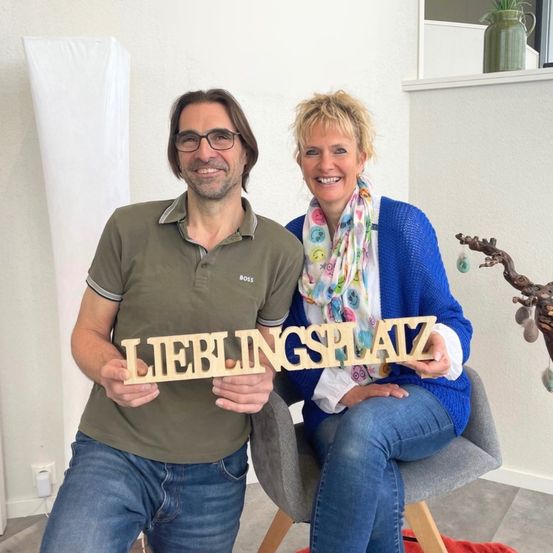 Ein Mann und eine Frau sitzen auf einem Stuhl in einem Zimmer, lächeln und halten ein Holzschild mit der Aufschrift 'Lieblingsplatz'.