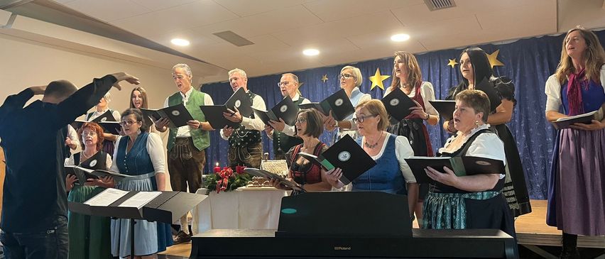 Ein Chor tritt auf der Bühne auf und singt mit Notenbüchern. Ein Klavier steht auf der Bühne und ein Notenständer daneben. Die Chormitglieder sind in traditioneller Kleidung gekleidet. Ein Tisch ist mit einem weißen Tuch bedeckt und hat Blumen und Kerzen darauf.