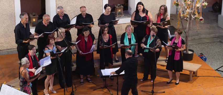 Ein Chor von Menschen singt auf der Bühne zusammen, mit einem Dirigenten an der Spitze. Sie halten Musikblätter in den Händen.