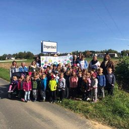 Eine Gruppe von Kindern und Erwachsenen posiert für ein Foto am Straßenrand mit einem Schild, das 'Stegersbach' besagt. Sie halten ein Banner mit dem Text 'Für die Zukunft'.