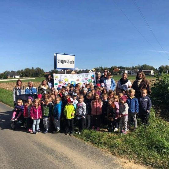Eine Gruppe von Kindern und Erwachsenen posiert für ein Foto am Straßenrand mit einem Schild, das 'Stegersbach' besagt. Sie halten ein Banner mit dem Text 'Für die Zukunft'.