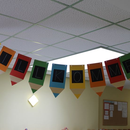 Ein farbenfrohes Banner aus Stiften hängt von der Decke und schreibt 'WILKOM'. Die Decke ist weiß mit einem quadratischen Licht. Eine Pinnwand mit Papieren ist an der Wand.