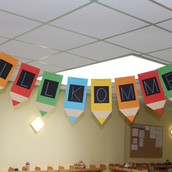 Eine farbenfrohe Klassenzimmerdekoration mit einem Banner, das 'Willkommen' sagt. Das Banner besteht aus farbigen Stiften und hängt an einer Decke mit einer quadratischen Leuchte.