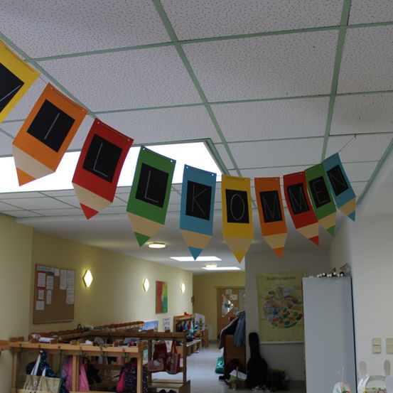Ein Flur mit bunten Bannern, die 'WELCOME' buchstabieren, hängt von einer gesprenkelten Decke. Verschiedene Gegenstände wie Taschen und eine Gitarre sind auf dem Boden verstreut.
