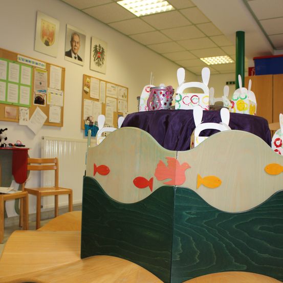 Ein Klassenzimmer mit Holzmöbeln und einer Anschlagtafel. Ein Tisch ist mit einem lila Tuch bedeckt, und Papierhasen und Fische sind darauf. Eine Anschlagtafel hat Papiere, die angeheftet sind.