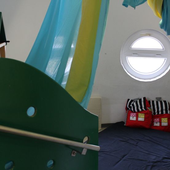 Ein kleiner Raum mit einem grünen Bett und einem runden Fenster. Bunte Kissen liegen auf dem Bett und bunte Vorhänge hängen an der Wand.