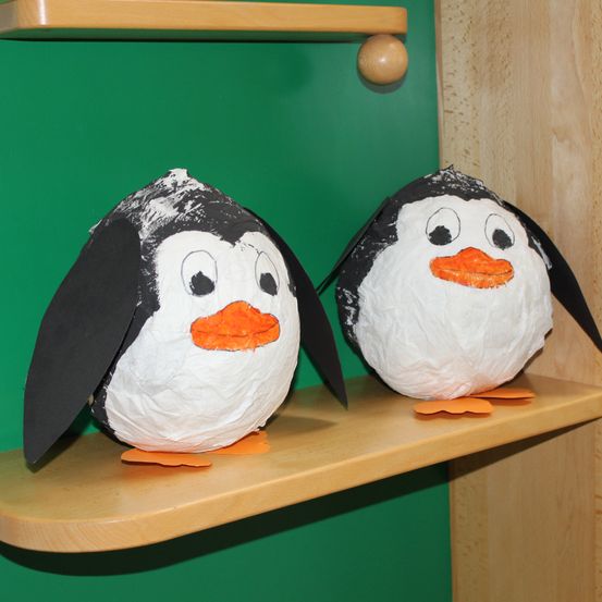 Zwei Pinguin-Kunstwerke mit schwarzen Flügeln, weißen Körpern und orangefarbenen Schnäbeln stehen auf einem Holzregal vor grünem Hintergrund.