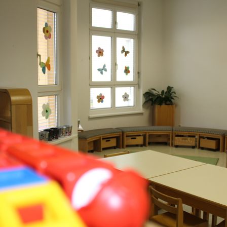 Ein Klassenzimmer mit weißen Wänden und zwei Fenstern mit Blumen- und Schmetterlingsaufklebern. Eine Bank mit Schubladen steht an der Wand, und eine Topfpflanze steht in der Ecke. Ein rotes Spielzeug im Vordergrund verdeckt einen Teil der Ansicht.