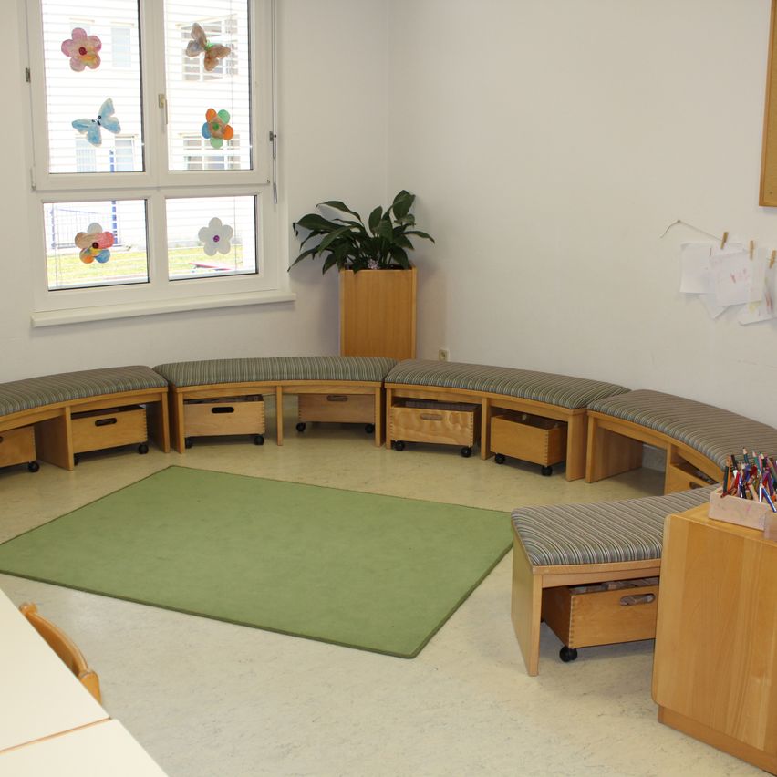 Ein Klassenzimmer mit weißen Wänden und einem großen Fenster mit bunten Blumenaufklebern. Ein grüner Teppich und Holzbänke umgeben eine Pflanze. Ein Holzlager mit einem Bleistiftkasten befindet sich rechts.