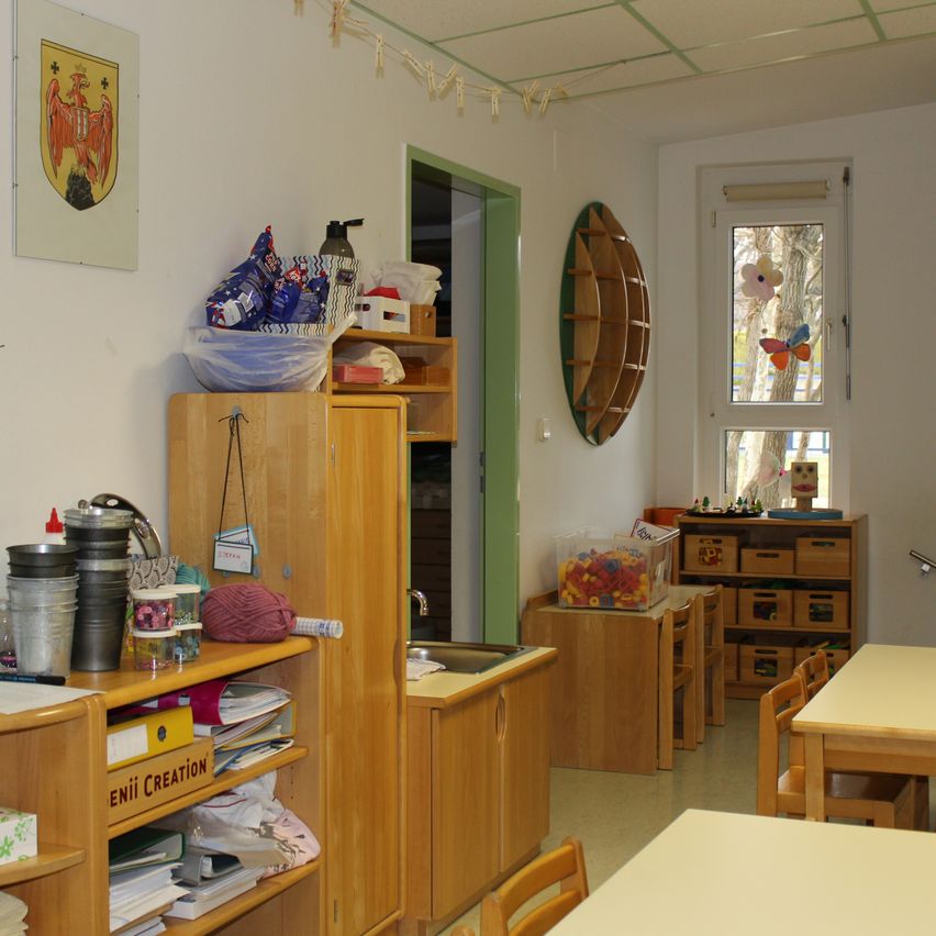 Ein kleiner Klassenraum mit hölzernen Tischen und Stühlen. Ein hölzerner Schrank hat Bücher und andere Gegenstände darauf. Ein Regal hat eine Flasche und andere Objekte. Eine Wand hat ein Schild mit einem Schmetterling.