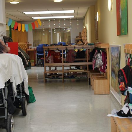 Ein leerer Flur mit gelben Wänden, Leuchtstofflampen und bunten Dekorationen. Holzbänke sind an der Wand angeordnet, mit Schuhen und Taschen darauf. Ein Kinderwagen ist mit einem weißen Tuch zu sehen.