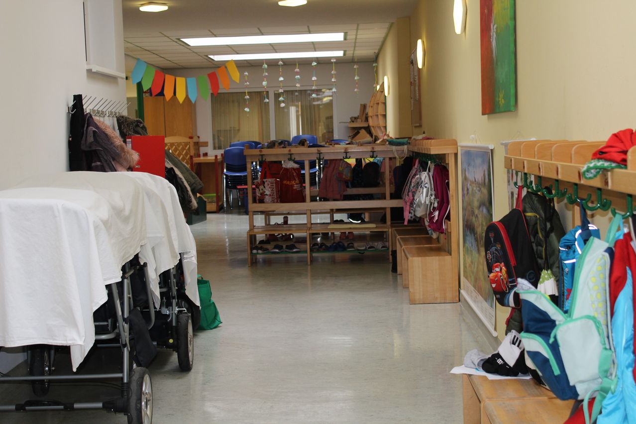 Ein leerer Flur mit gelben Wänden, Leuchtstofflampen und bunten Dekorationen. Holzbänke sind an der Wand angeordnet, mit Schuhen und Taschen darauf. Ein Kinderwagen ist mit einem weißen Tuch zu sehen.