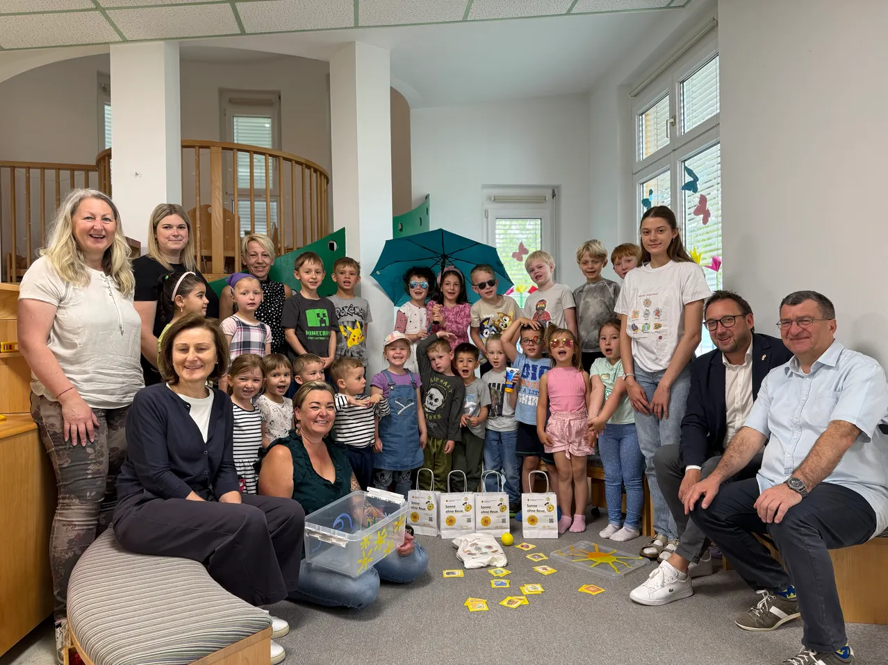 Eine Gruppe von Erwachsenen und Kindern, darunter eine mit Hut, steht und sitzt in einem Raum. Einige von ihnen tragen Brillen und alle lächeln. Auf dem Boden befinden sich mehrere gelbe Objekte, möglicherweise ein Spielzeug, und einige Taschen.