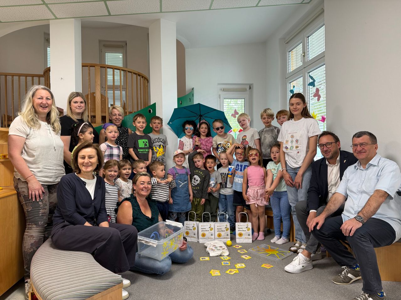 Eine Gruppe von Erwachsenen und Kindern, darunter eine mit Hut, steht und sitzt in einem Raum. Einige von ihnen tragen Brillen und alle lächeln. Auf dem Boden befinden sich mehrere gelbe Objekte, möglicherweise ein Spielzeug, und einige Taschen.